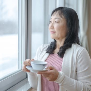 窓の外を眺めながら飲み物を飲む中年女性のイメージ画像