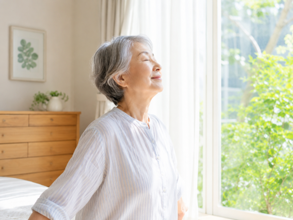 朝窓辺で深呼吸をしているシニア女性のイメージ画像